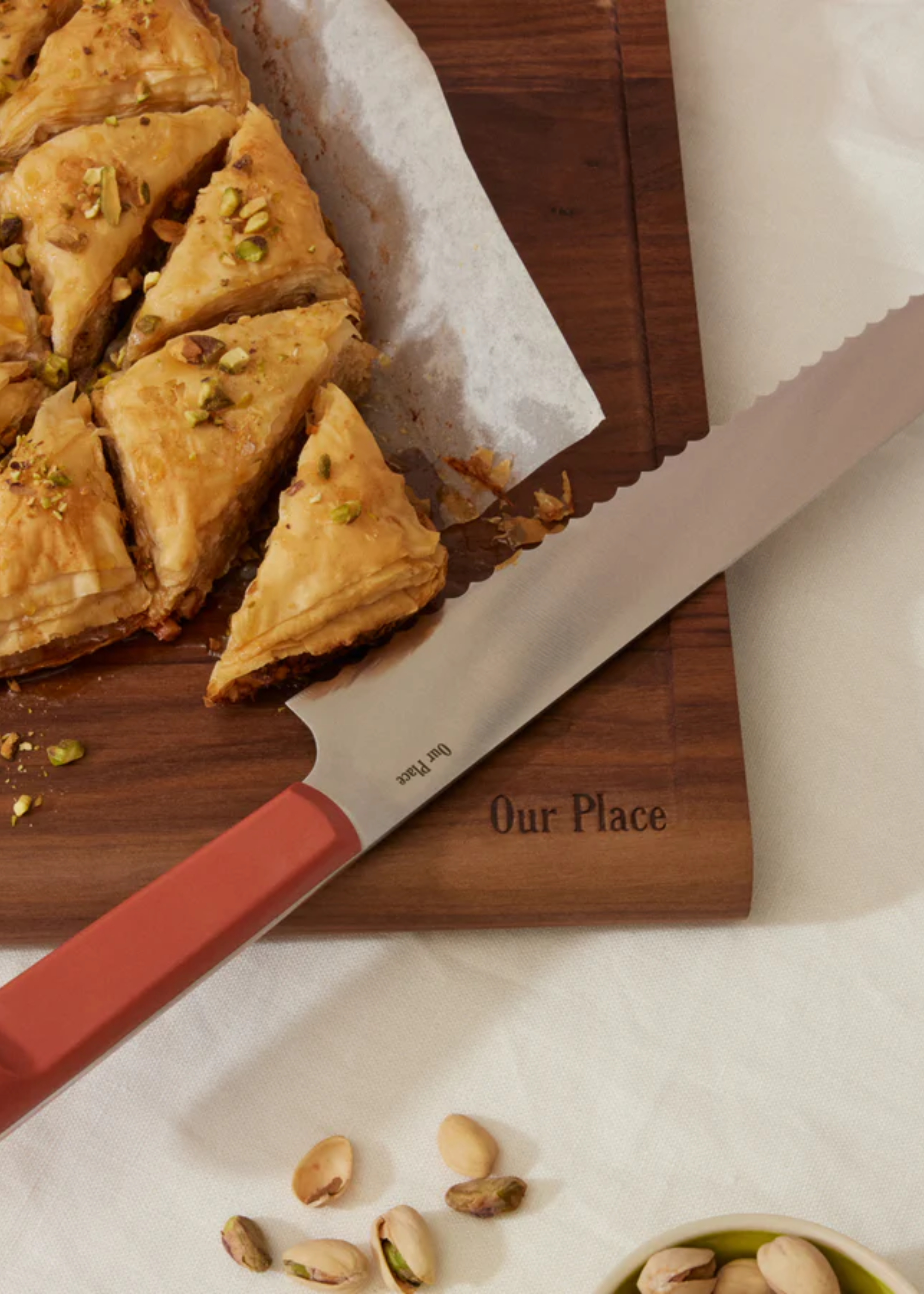 A serrated knife with a pinkish red handle near triangled baklava on a baking sheet on a wood chopping board