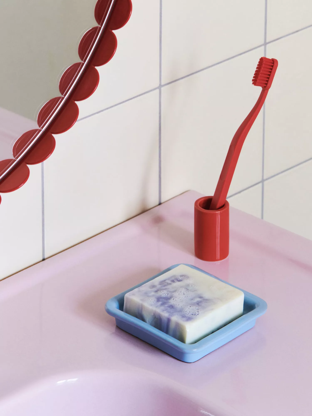 A cubed handmade soap with lavender from HAY on a bathroom sink counter beside a red toothbrush and mirror