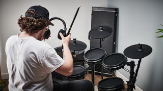 Drummer sat at an e-kit places a pair of headphones over his head