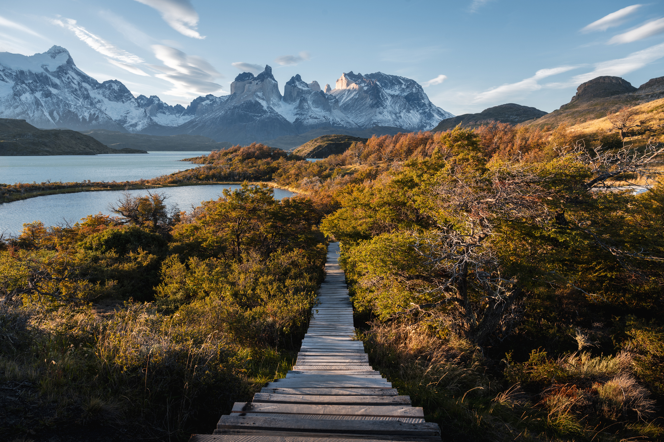 a wooden plank walking path leading to a lake surrounded by snow-capped mountains