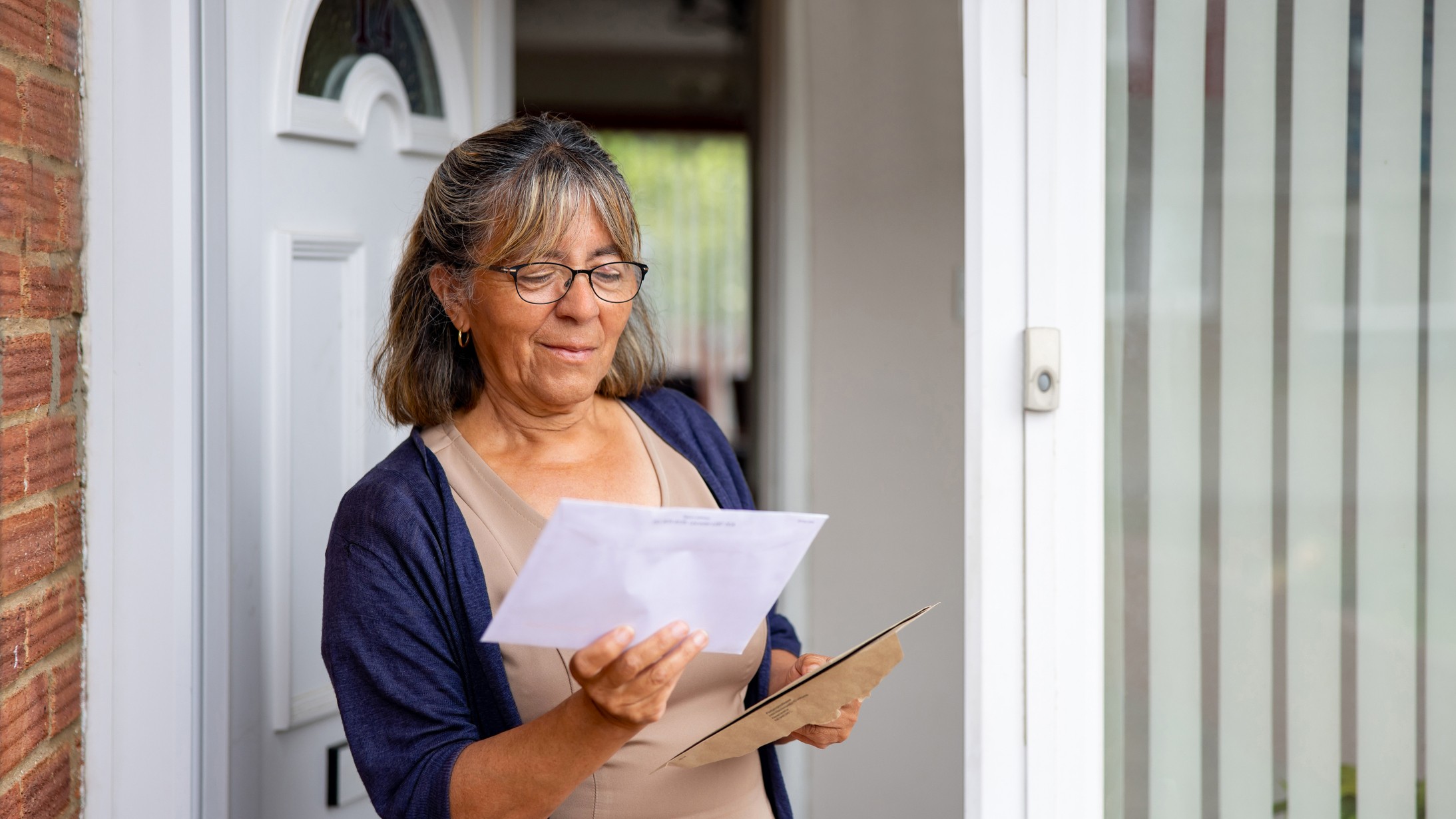 Mature woman at the front door of her house checking her mail and reading an envelope.