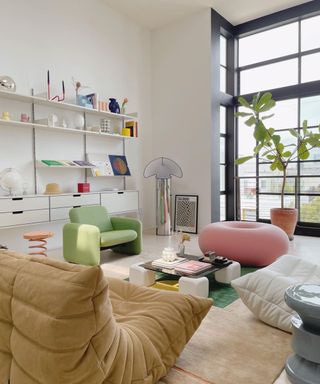 A small living room with white walls, colorful seating, wall shelving with decor on it, and a large window