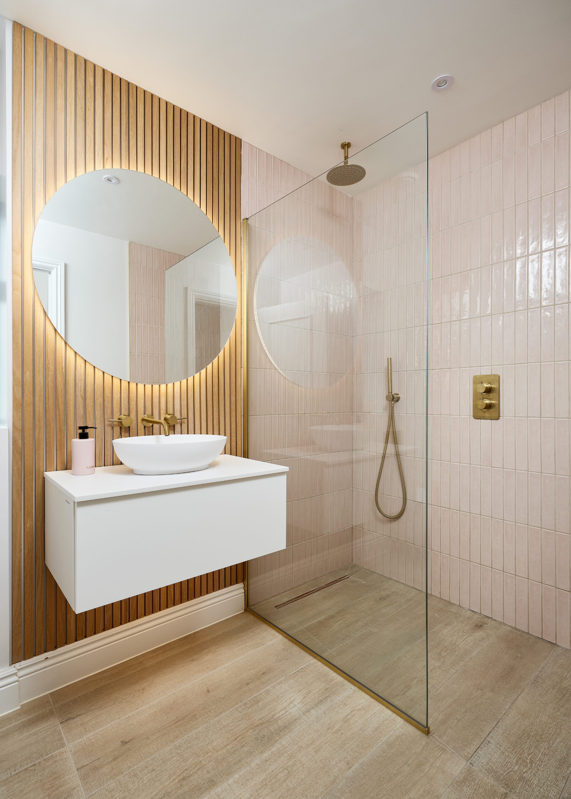 A sleek modern shower with a barely there frame and a wall-hung white vanity unit with basin on top and an oversized round mirror with a backlit glow. These are all mounted to a fluted wood wall.