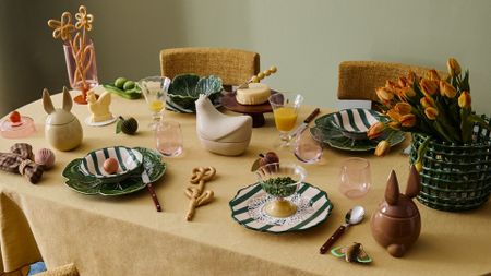 A table set for Easter with bunny-eared jars, stripey plates, a woven ceramic basket with yellow tulips, cabbageware, and cutlery