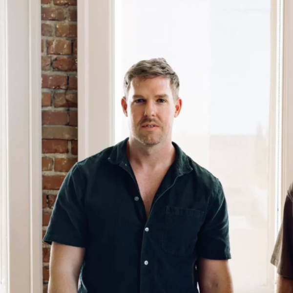 A headshot of Nate Fox: a man staring at the camera with medium-toned brown hair swept slightly to one side, wearing a black short-sleeved shirt with a couple of buttons undone in front of large white-framed windows within a brick wall