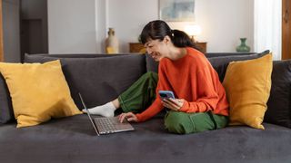 Woman sitting on sofa, holding phone and looking at laptop