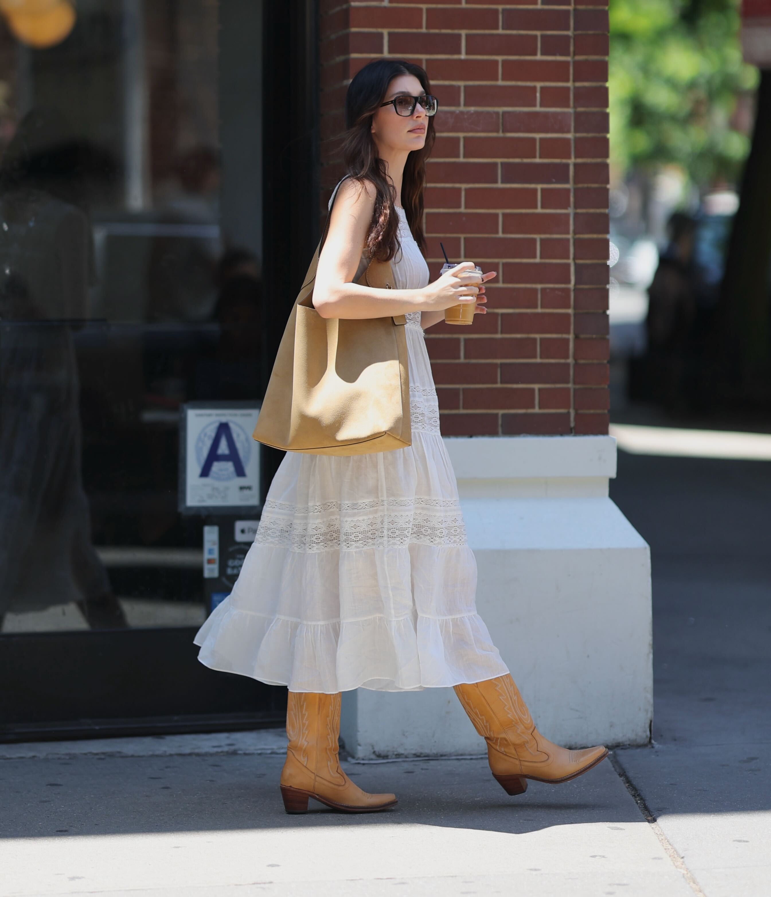 Camila Morrone wears a coach bag, white sundress, and tan cowboy boots.