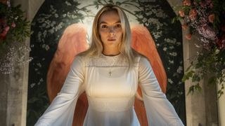Jodie Comer as Villanelle, in a white garment standing in front of a painting of angel wings, in Killing Eve season 4 episode 1 