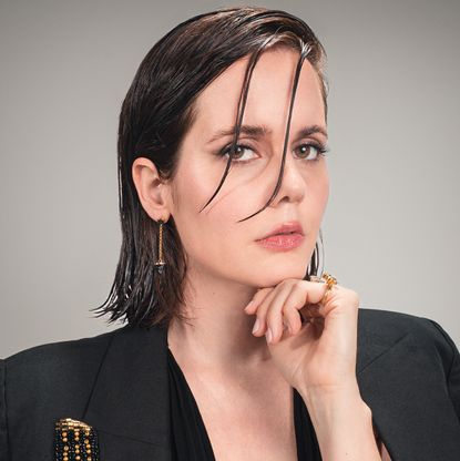 actress ashley sutton wears a black blazer and poses with her hand under her chin and two strands of hair in front of her face