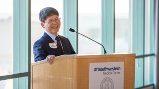 older man with glasses wears a blue suit and red tie while smiling at a podium with the words "ut southwestern medical center" on the front