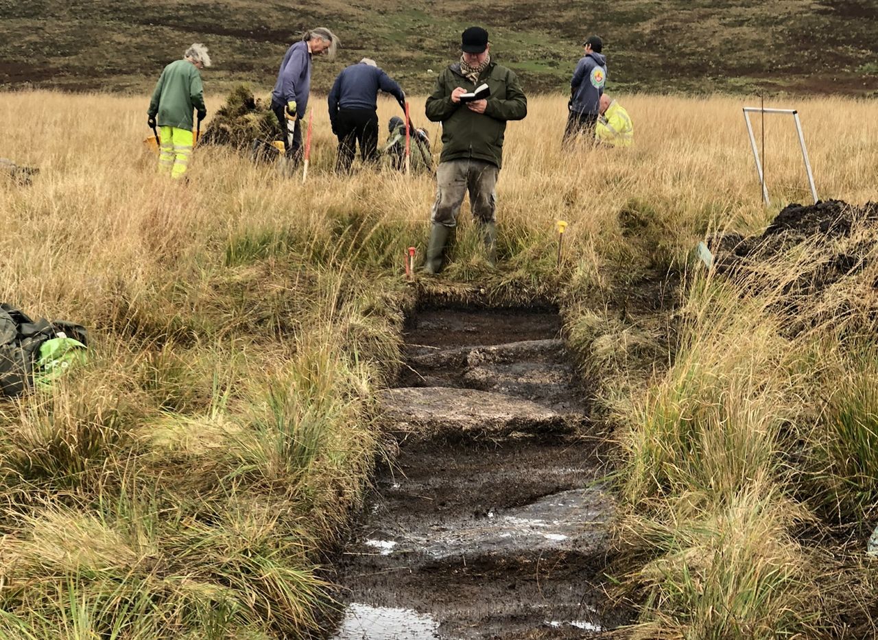 A group of people at an excavation site behind a rectangular dirt hole