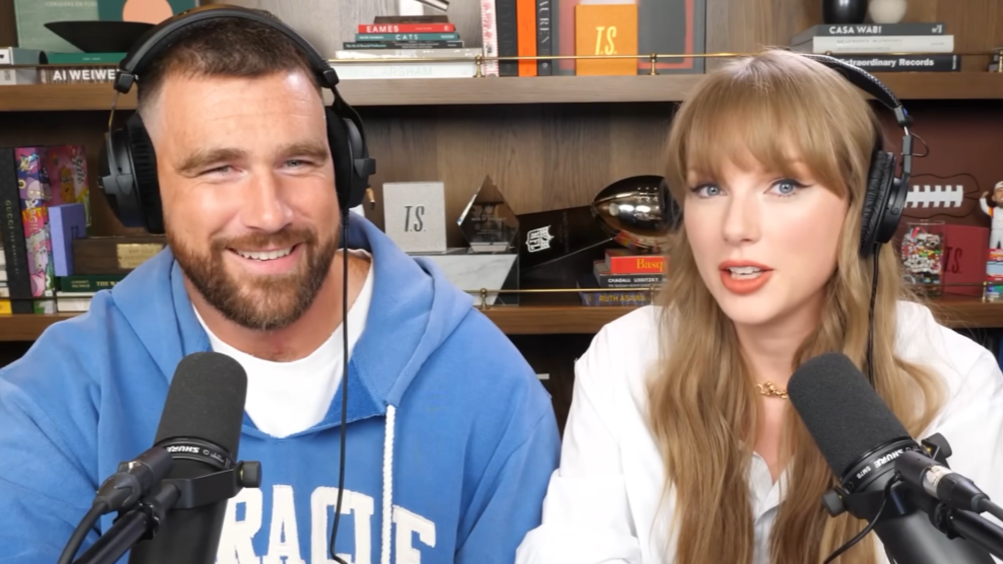 From left to right: Travis Kelce smiling and Taylor Swift smiling, they're both wearing headphones and talking into mics on New Heights. 