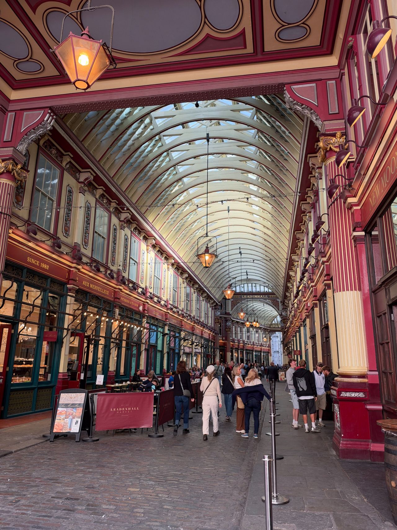 Interior of Leadenhall Market in London, with ornate red-and-cream architecture, hanging lamps, shops, and visitors.
