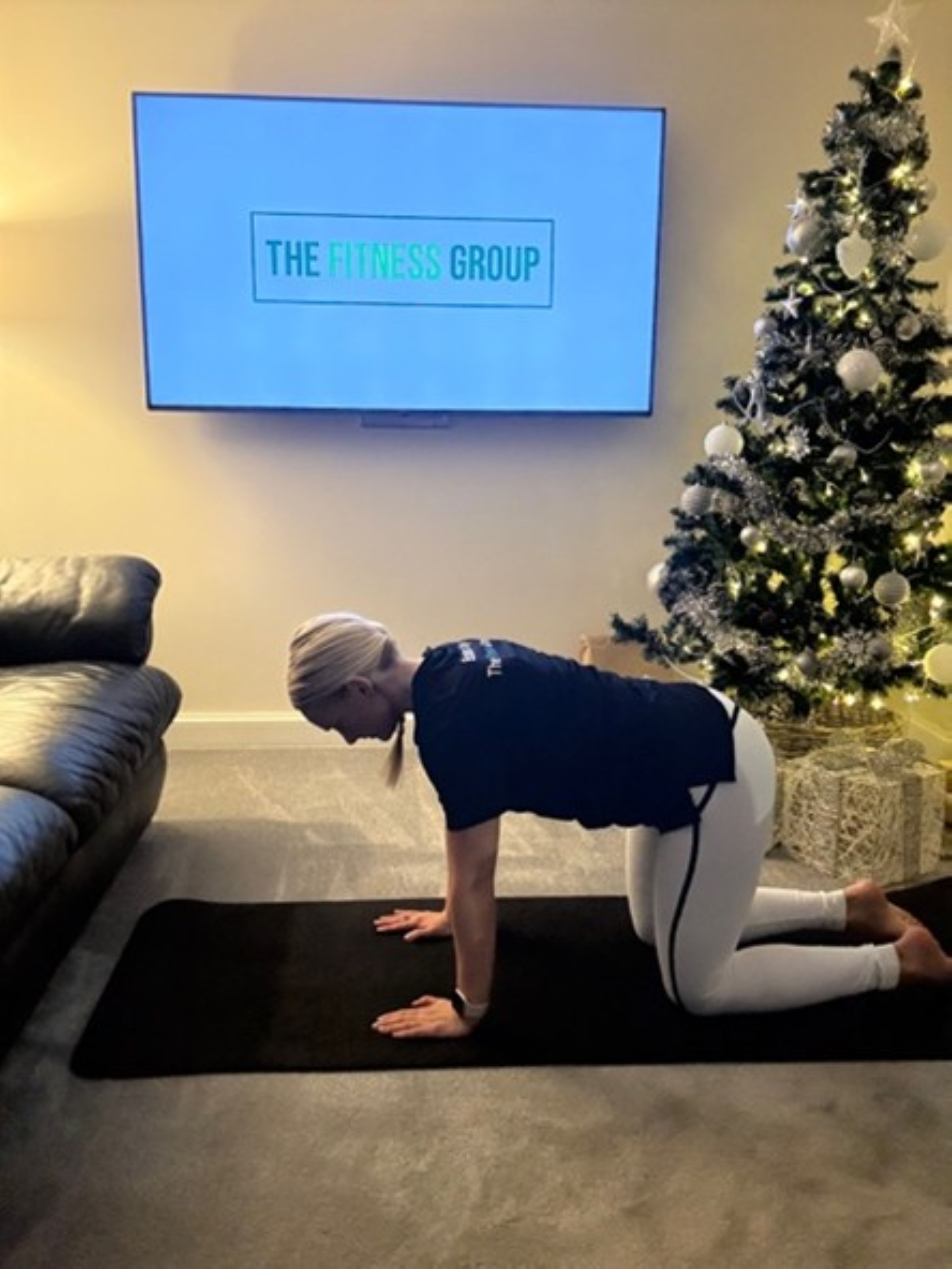 Woman demonstrates a Pilates exercise between a sofa and a Christmas tree. A mounted TV in the background reads "The Fitness Group"