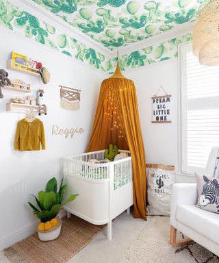 white nursery with botanical green wallpapered ceiling, mustard yellow canopy, wall art, rugs on floor with lounge chair