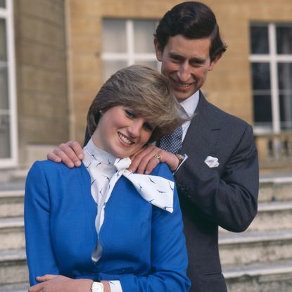 Prince Charles wearing a gray suit laughing and posing with his hands on Princess Diana's shoulders in front of a staircase in their engagement photos 