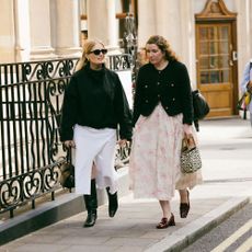 women walk the streets of london in fashionable outfits. one wears black sweater and white skirt while the other wears black cardigan sweater and fun pink skirt