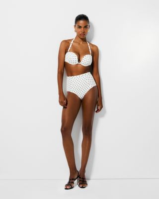 A model wears a white and black polka dot Reformation swimsuit in front of a plain backdrop