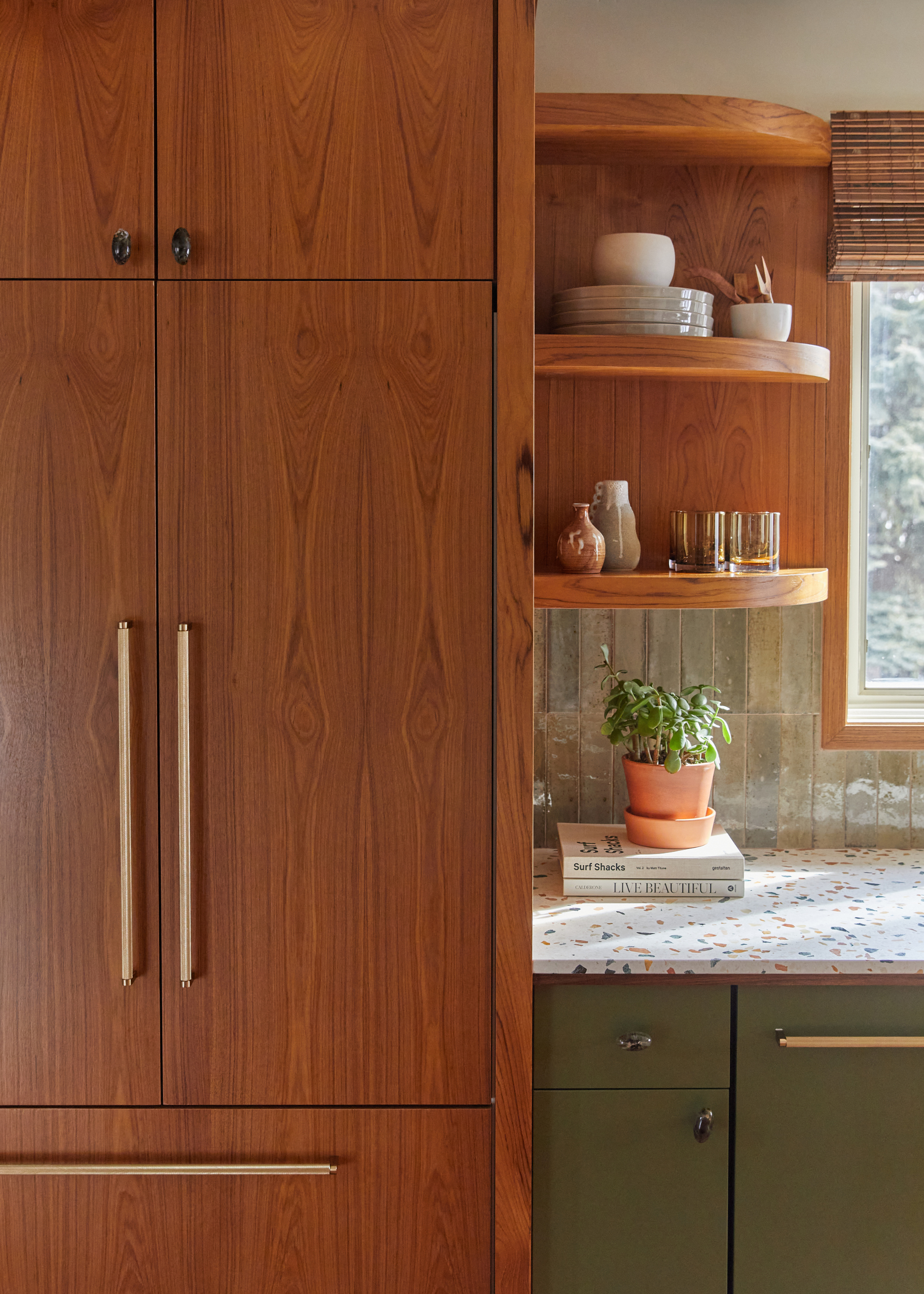A kitchen with wooden cabinets, brushed gold handles, a terrazzo counter, bowls, plates, and a potted plant
