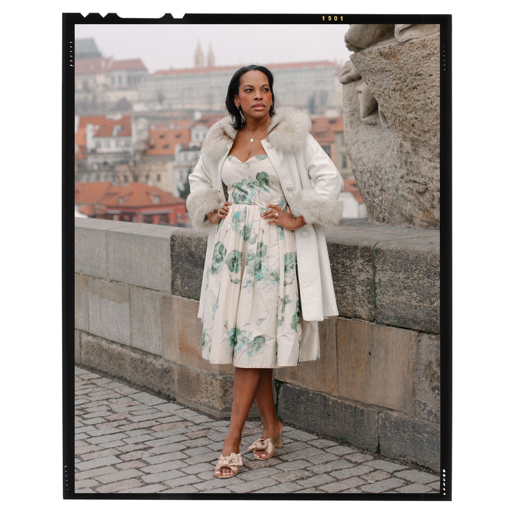 portrait of Sarah Kunst in a white and blue floral dress, white fur-trimmed coat, and tan bow heels standing on a cobblestone street
