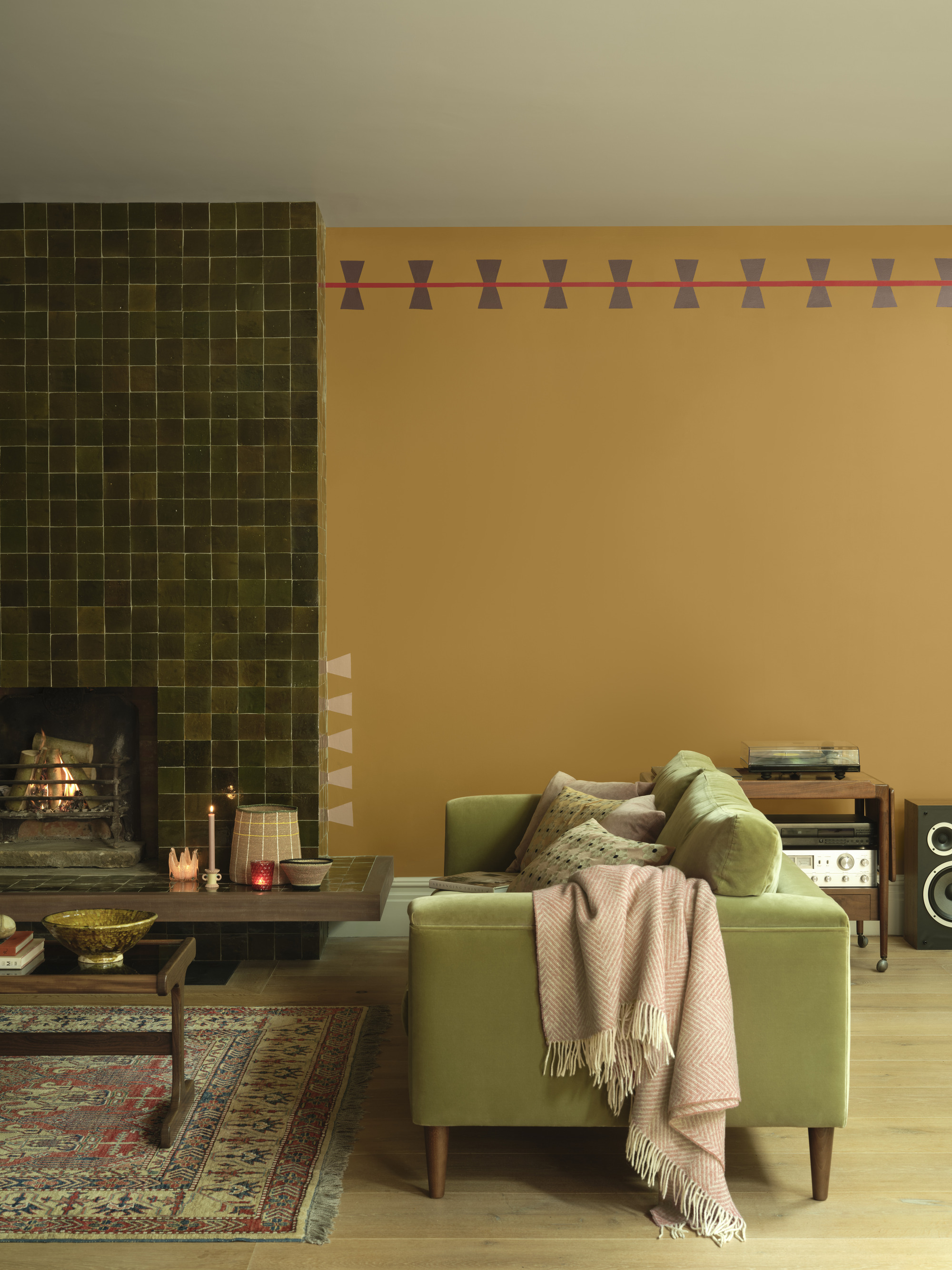 modern living room with golden yellow wall, painted trim detail, light green sofa with throw, timber floors, persian rug, and green tiled fireplace