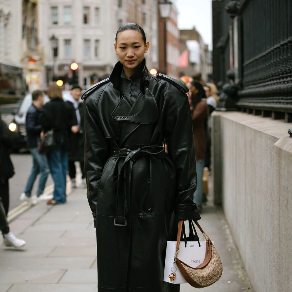 A woman poses in an all black outfit. Included in her look are a duster length leather trench coat and black adidas. 