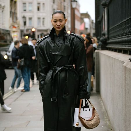 A woman poses in an all black outfit. Included in her look are a duster length leather trench coat and black adidas.