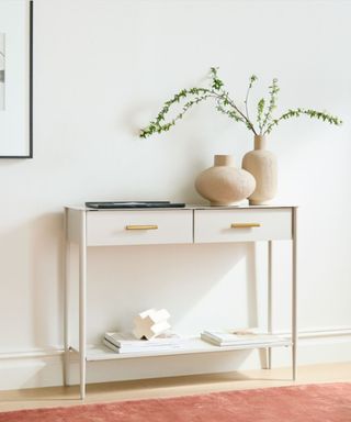 A white console table with a plant and vase on it next to a white wall, on top of wooden flooring with a red rug on it