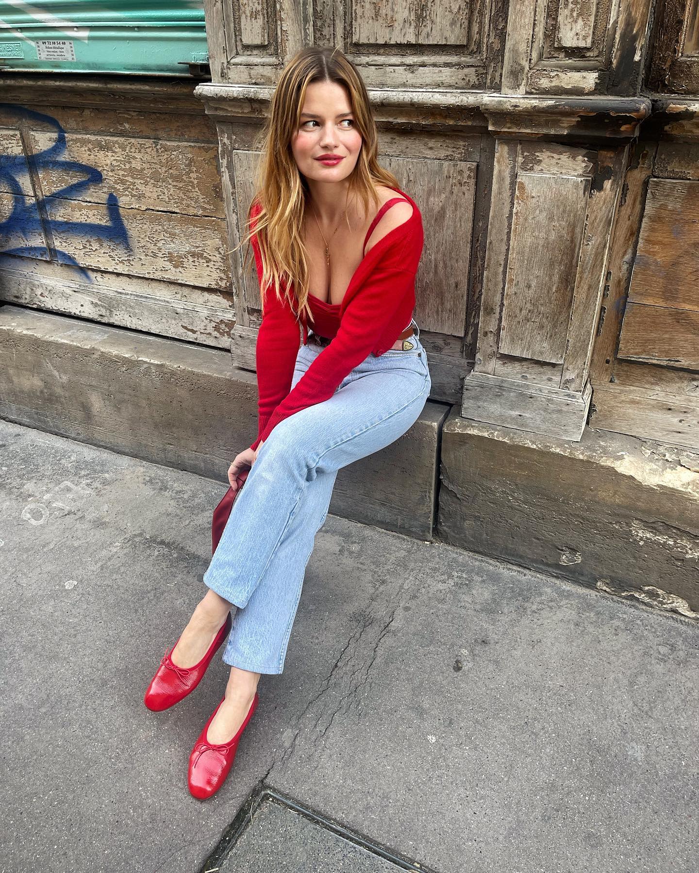 French influencer Sabina Socol sits on a ledge on a Paris sidewalk wearing a red cardigan, a red tank top, light-wash jeans, and red ballet flats