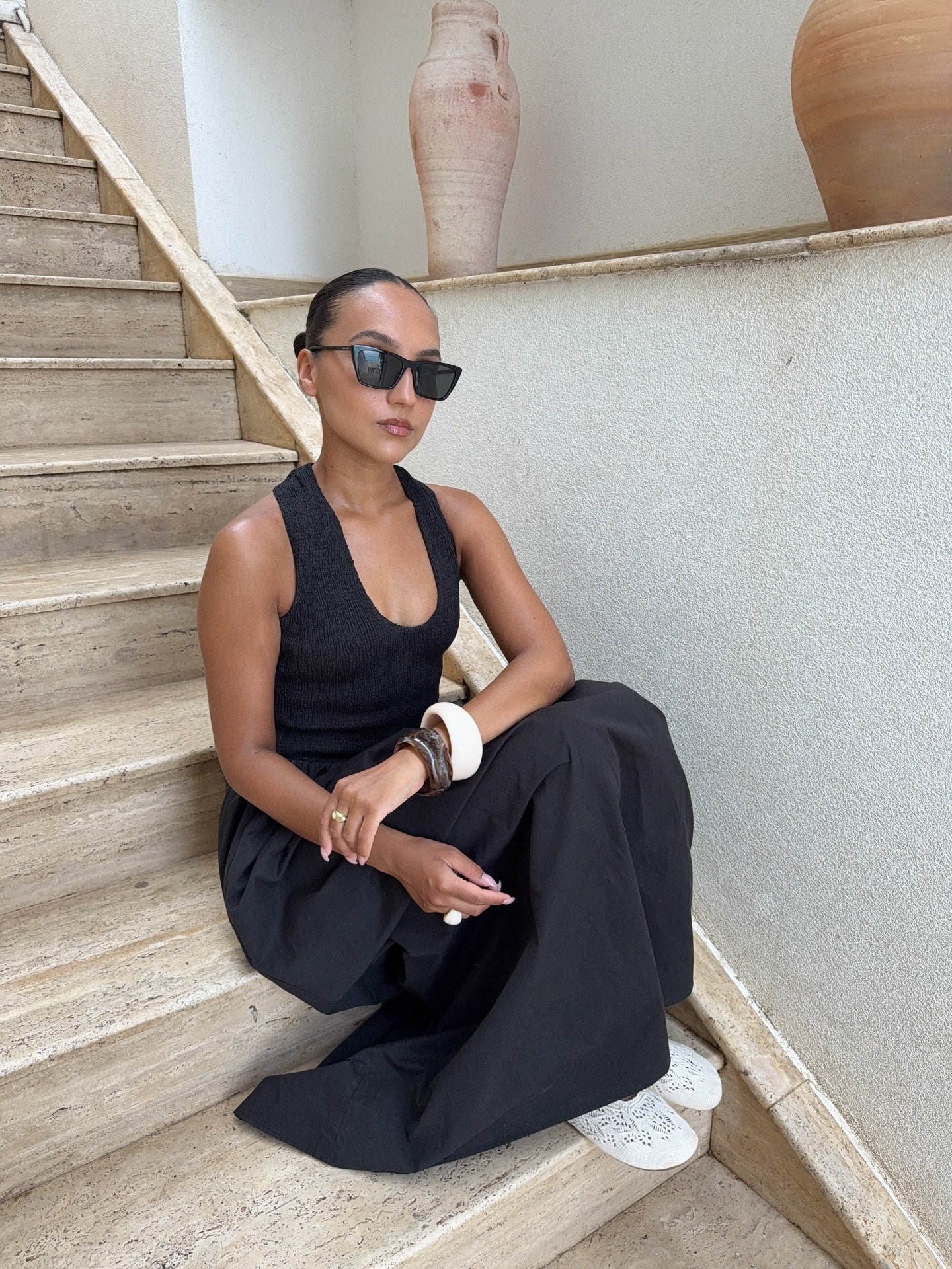 Fashion person Ingrid Edvinson wears a black halterneck maxi dress crocket ballet flats, ceramic bangles and cat eye glasses. She is sat on cream steps in the recent photo taken from her Instagram.