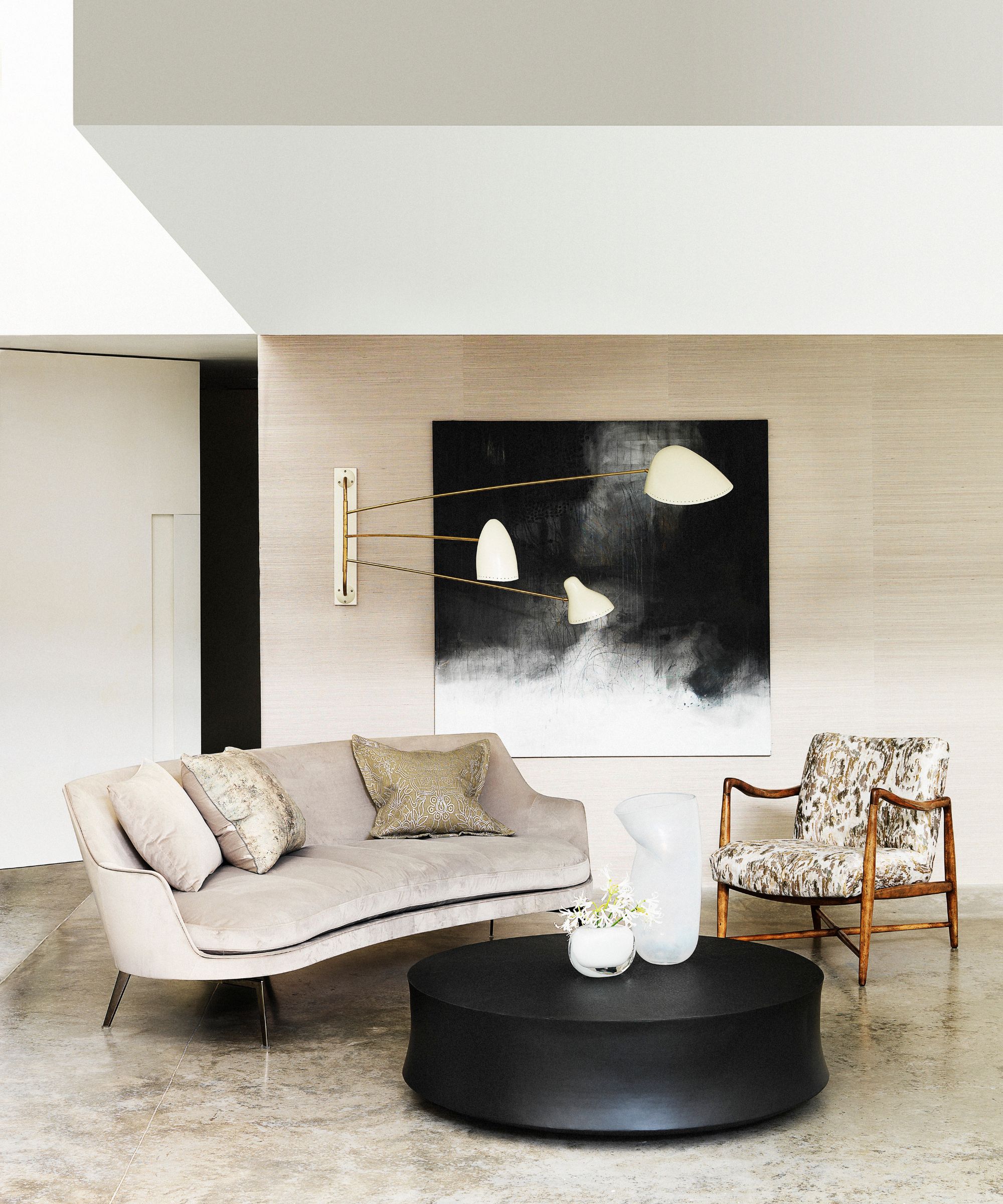 A neutral, minimalist living room with a grey couch with neutral cushions, an upholstered chair with wooden armrests, and a large black circular coffee table with a plant and white vase on top. On the beige wall behind is a large black artwork.