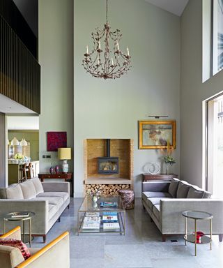 double height living room with gray green walls, chandelier, woodburner, neutral sofas and armchair