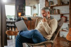 A mature man is sitting at home with his laptop in a chair focusing on his work.