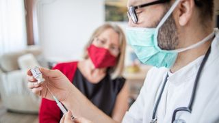 A man in a surgical mask and gloves preps a COVID-19 vaccine for a masked patient who's sitting in the background