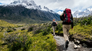 A pair of hikers are moving towards the mountains, they carry packs and are dressed for cooler weather in pants and jackets. 