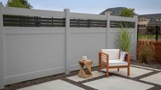 A wooden patio chair with white cushions, throw, twisted design side table on paver patio with a tall gray fence behind 