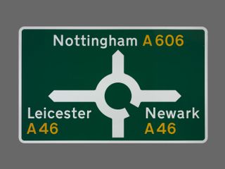 A road sign designed by Margaret Calvert and Jock Kinneir. The green background of the sign features a roundabout symbol with words, Nottingham A606, Leicester A46, Newark A46, written in white and yellow type