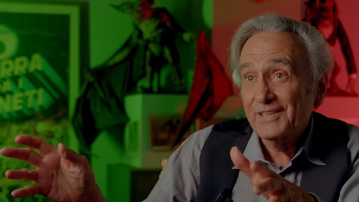 Filmmaker Joe Dante animatedly discusses Hammer Horror movies in a red-and-green lit room filled with Gremlins props for Ben Field&#039;s feature documentary 