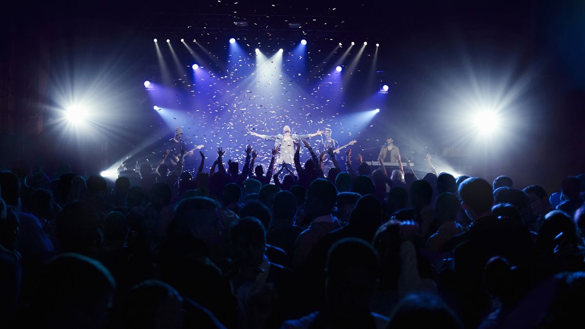 A lead singer stands arms spread in front of a raucous concert crowd.