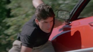 Charlie Sheen walking and ducking down next to a red car in Navy SEALs