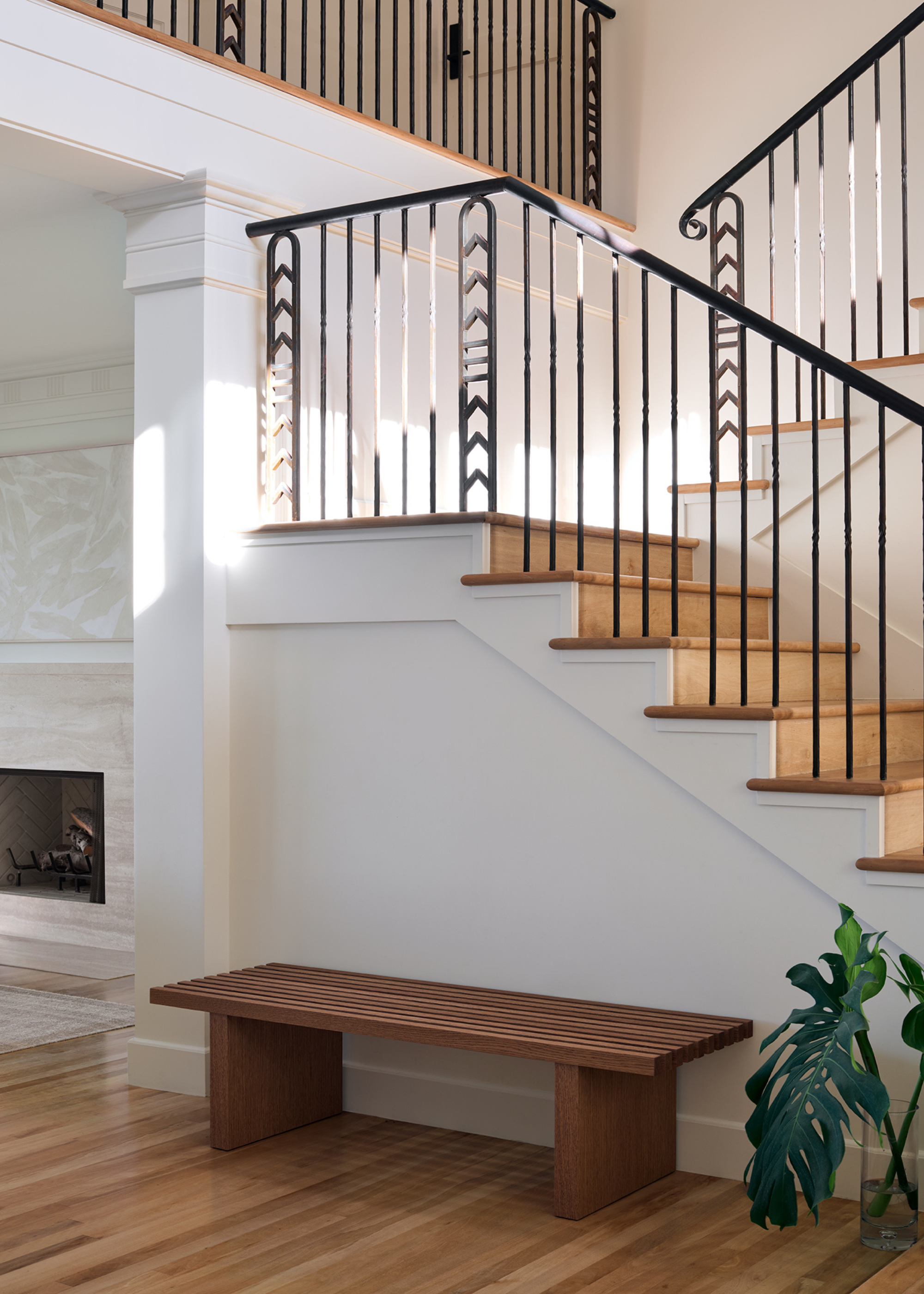 An airy entryway by a wood staircase with a wood bench, a vase of cut leaves and wood flooring