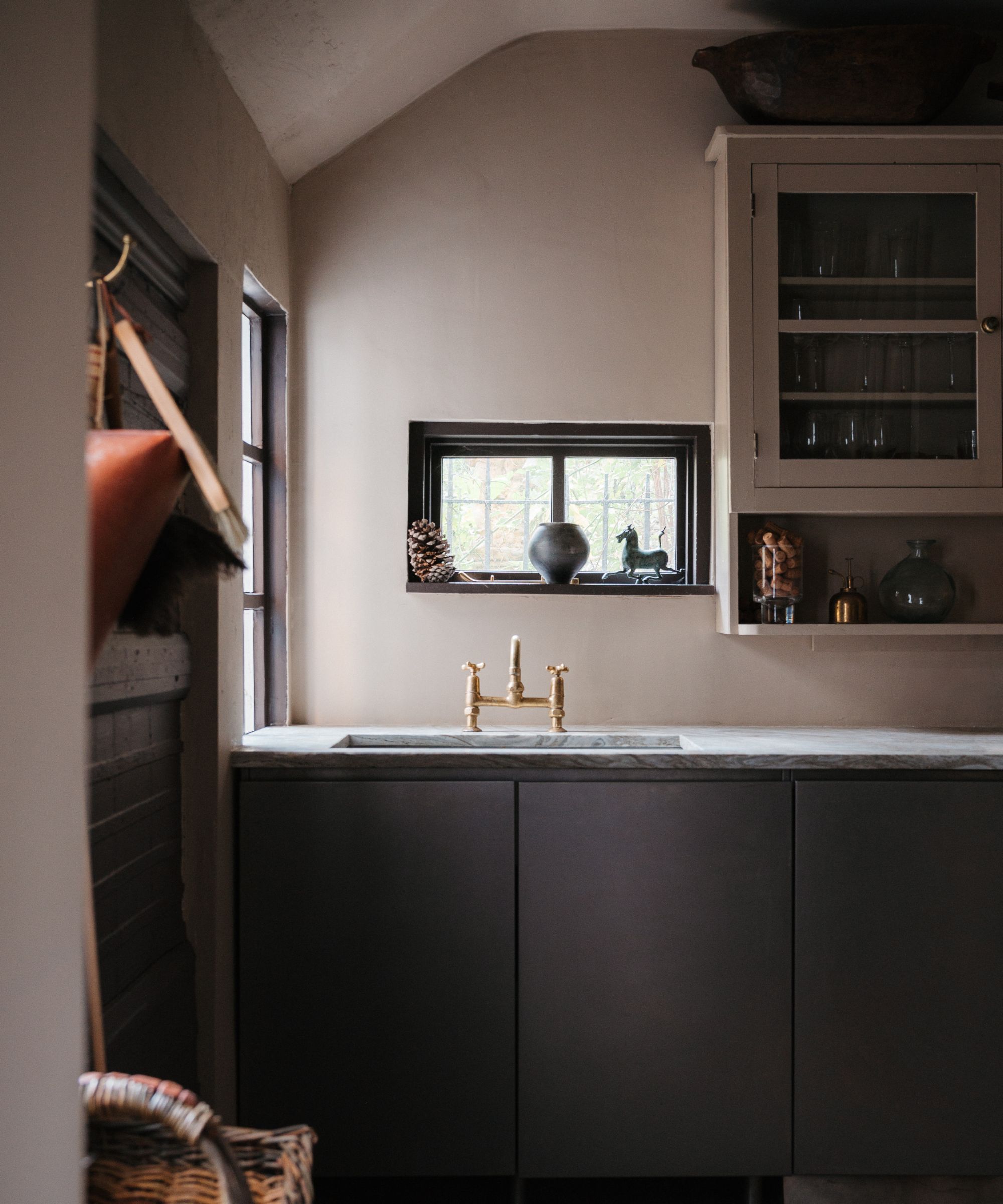 Moody kitchen featuring dark charcoal cabinets, a light marble countertop, a brass faucet, and a small window displaying a pinecone and decorative objects