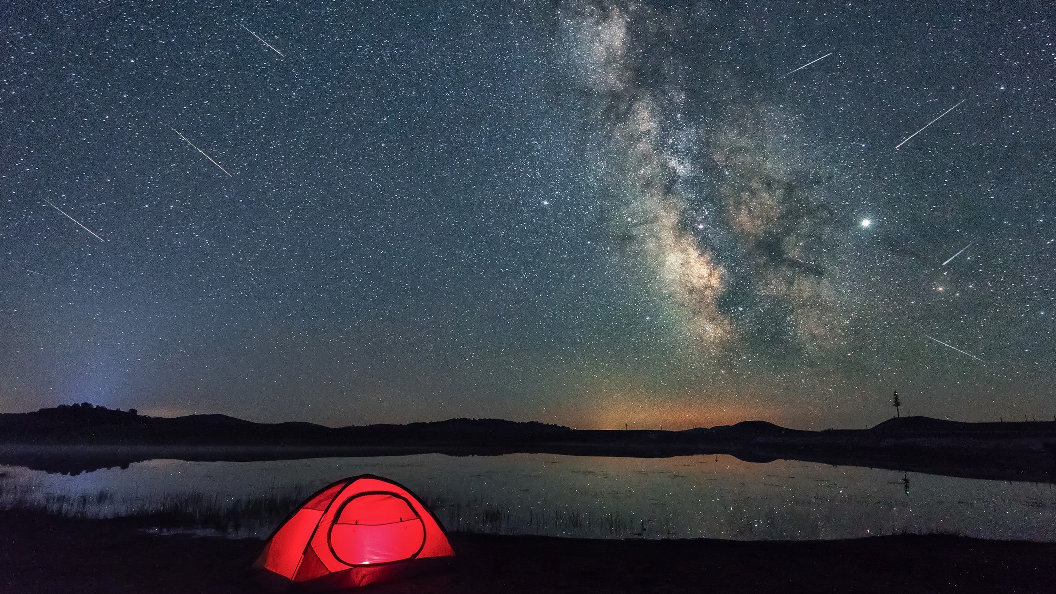 streaks of perseid meteors in the sky against the milky way backdrop and a small red tent in the foreground.