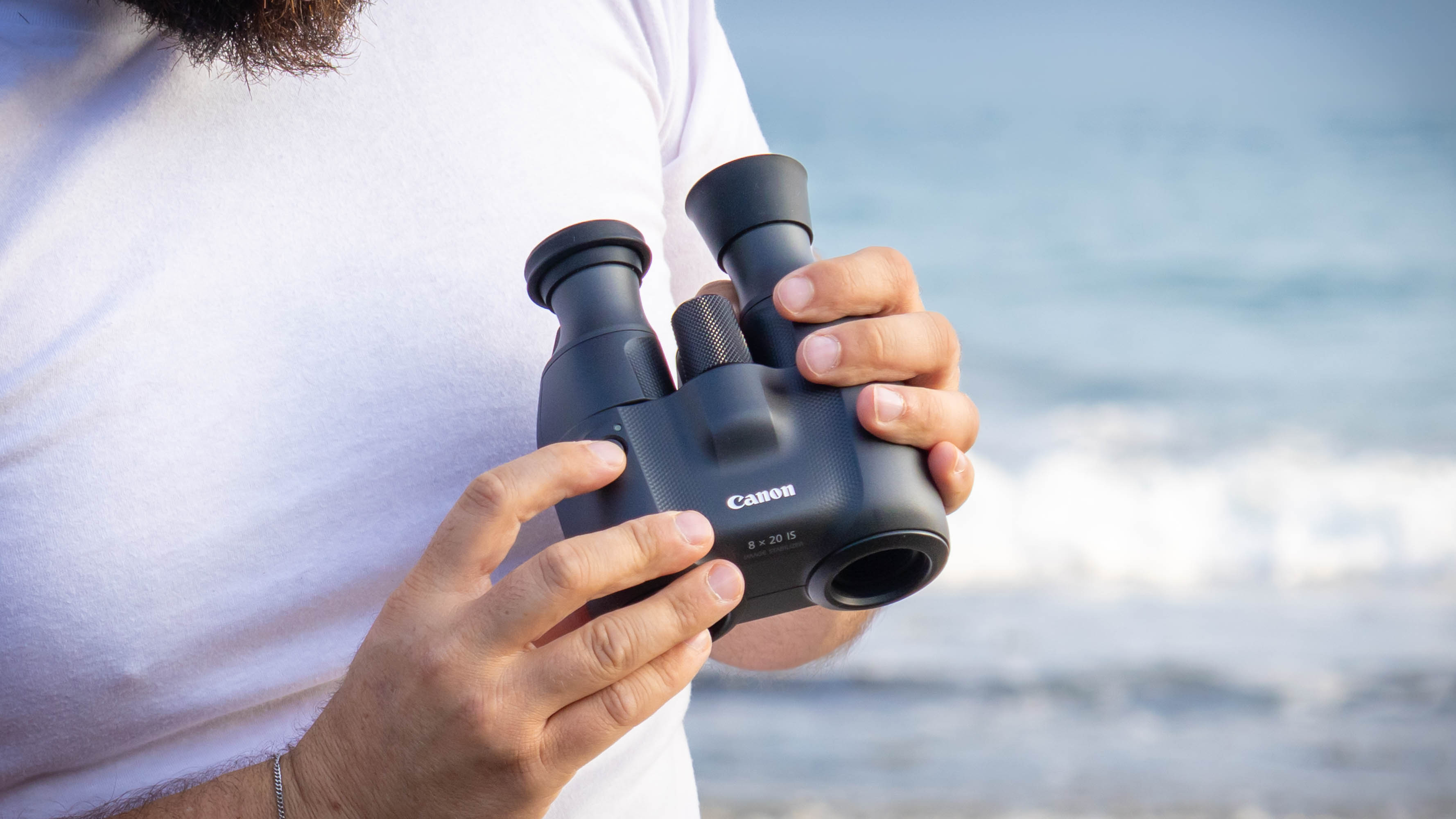 A person holding the Canon 8x20 IS binoculars with one eyecup flipped up and their finger over the IS button.