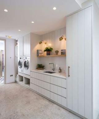 Bright and white laundry room with washer-dryer built into cabinets, potted plants, utility sink, and brass task wall lights