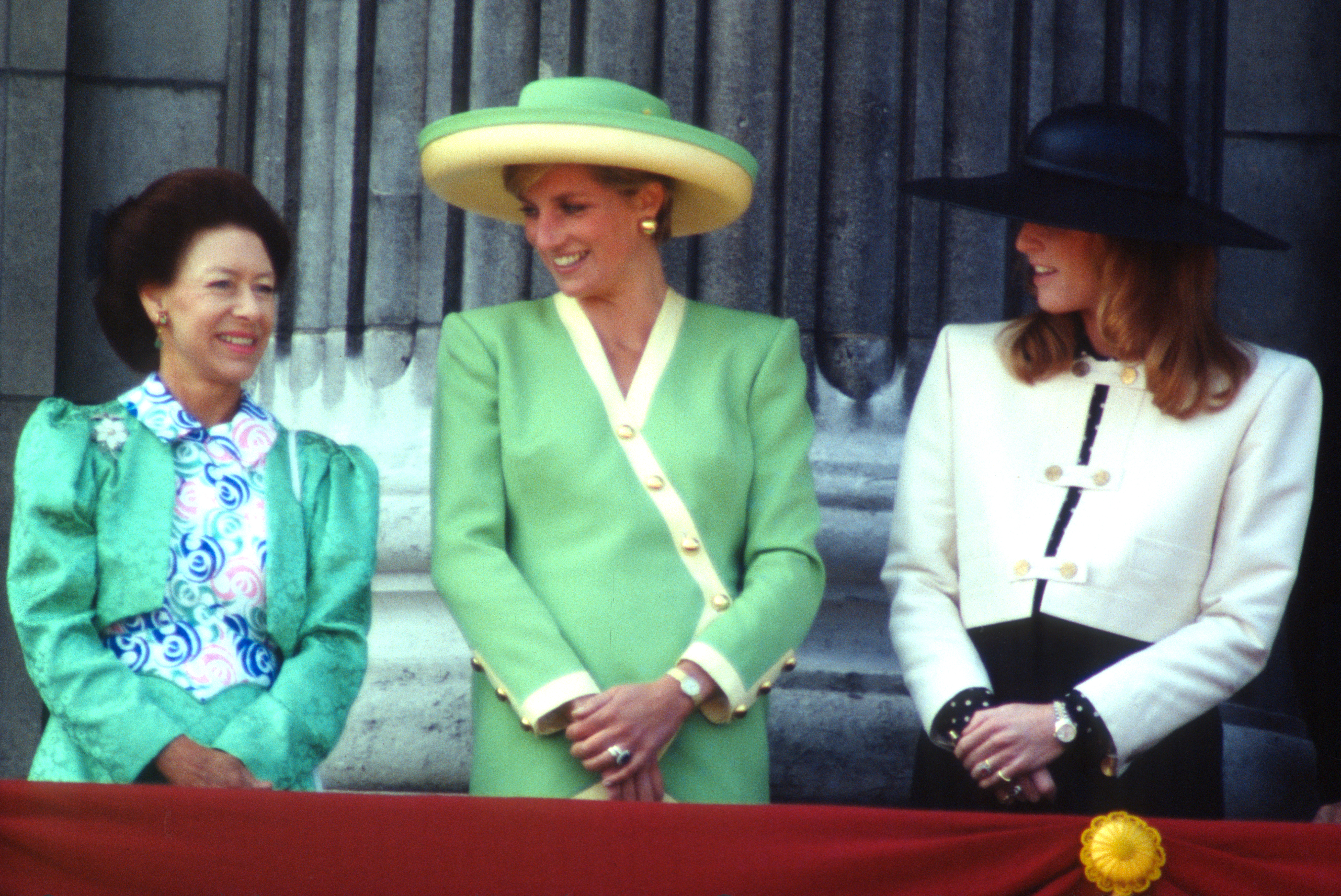 Princess Margaret wearing a green dress smiling and talking to Princess Diana, wearing a green suit and hat, and Sarah Ferguson wearing a white jacket