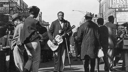 A black and white street photograph of a man standing in the middle of a crowded Maxwell Street Market holding a resonator guitar.