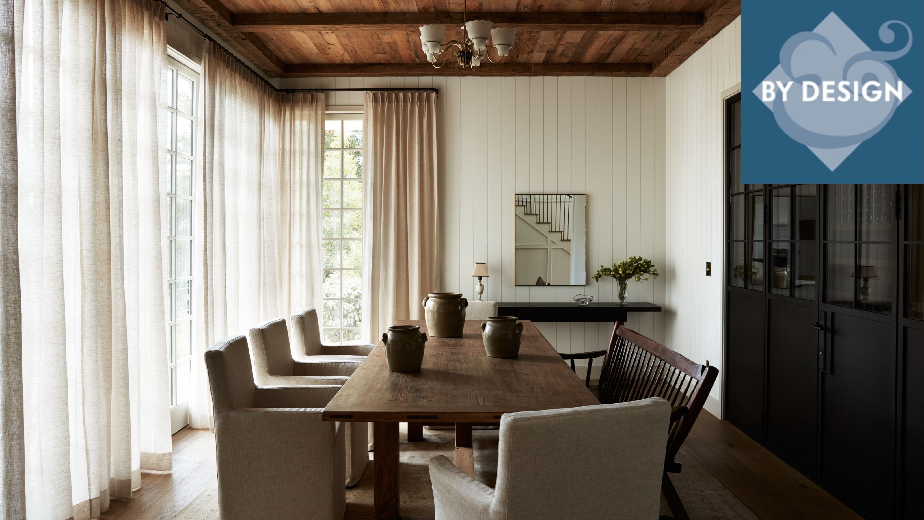 Neutral dining room with white panelled walls, long dark wood dining table with upholstered chairs and bench, sheer curtains on windows, black cabinetry