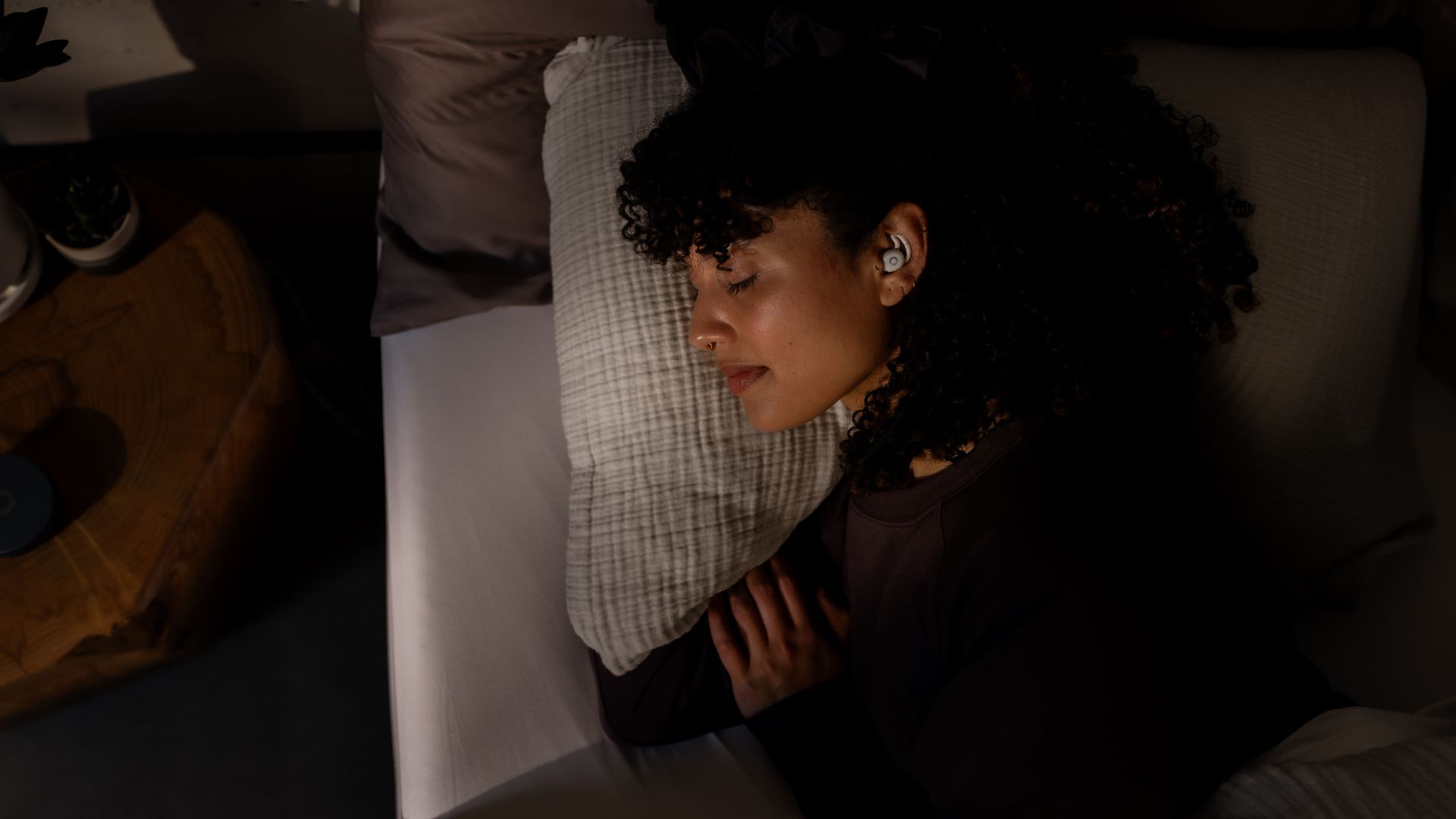 A woman with dark curly hair sleeps on her side with a white pair of Ozlo Sleepbuds 2 visible in her ear