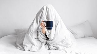 A person wrapped up in a blanket, sitting on a bed, holding a cup of coffee